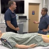 Two individuals in a medical training room are discussing near a hospital bed with a medical training mannequin lying on it. The room contains medical equipment and monitors.