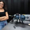 A person with curly hair and glasses stands smiling next to a laboratory setup involving lasers and optical equipment. 