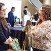 Two people are engaged in conversation at an indoor event. They stand near a table with chairs, and several other people are interacting in the background. The setting appears to be professional.