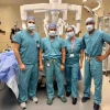 Four healthcare professionals in surgical attire stand in an operating room. They wear blue scrubs, masks, and caps. Behind them is a large piece of medical equipment. The room is well-equipped with medical supplies and monitors.