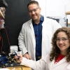 Two people in lab coats are in a laboratory setting, surrounded by electronic equipment and wires.