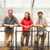 Three individuals stand inside a greenhouse, leaning on metal railings. 