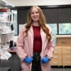 A person stands confidently in a laboratory setting, wearing a pink lab coat and blue gloves. The laboratory is filled with equipment, shelves, and supplies.