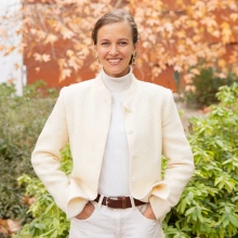 A person standing outdoors with a bright smile, wearing a white jacket, turtleneck, and trousers. The background shows trees with autumn leaves.