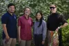 Four people smiling and standing together outdoors, with greenery in the background. From left to right, one person is wearing a blue polo shirt, the next is in a red button-down, followed by a person in a light blue shirt, and another in a black top with lace sleeves.